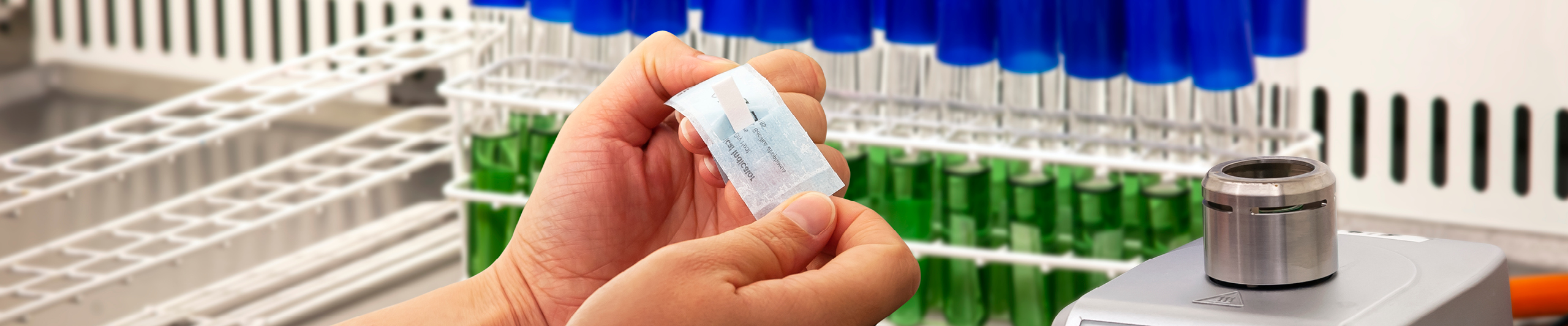Et billede af emballagen fra Steris med de biologiske indikatorer (Sporadi). I baggrunden ses reagensglas med blå hætter og grøn væske til bakteriesporing.